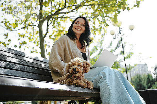 Lächelnder Freiberufler sitzt mit Laptop und Hund auf Bank im Park