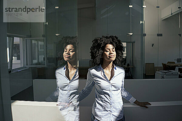 Geschäftsfrau mit geschlossenen Augen steht im Büro im Sonnenlicht