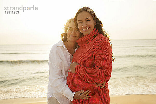 Glückliche Mutter umarmt schwangere Tochter am Strand