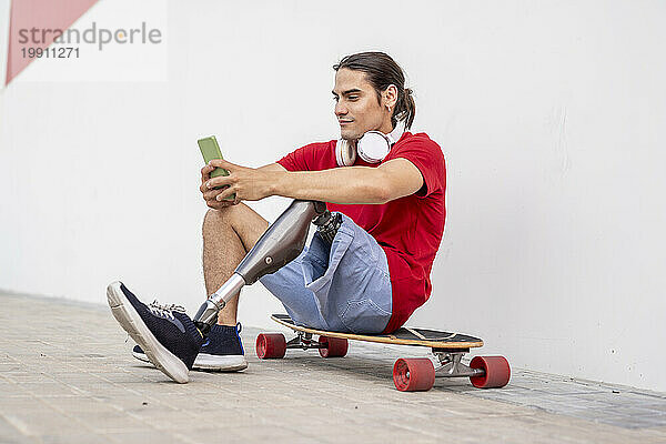 Lächelnder junger Mann sitzt auf Skateboard mit Beinprothese und benutzt Smartphone