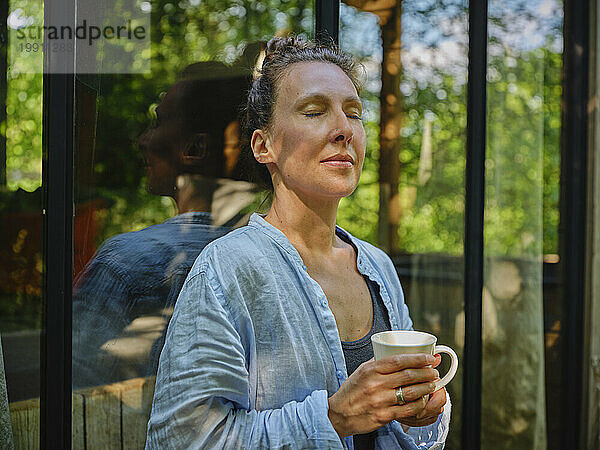 Frau mit geschlossenen Augen hält Kaffeetasse in der Hand und lehnt am Fenster