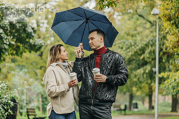 Paar hält Kaffeetassen und Regenschirm im Herbstpark