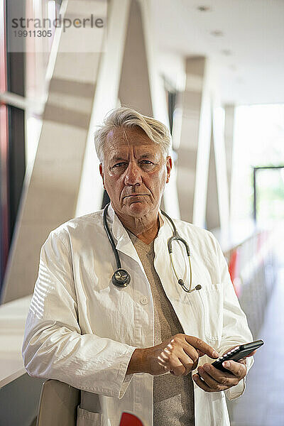 Ernsthafter Oberarzt steht mit Smartphone