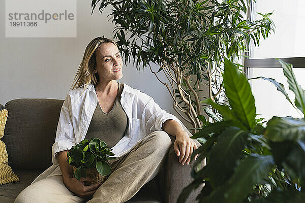 Nachdenkliche blonde Frau mit Topfpflanze sitzt zu Hause auf dem Sofa