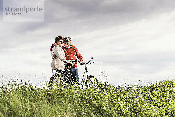 Glückliches älteres Paar  das mit dem Fahrrad auf dem Feld unter bewölktem Himmel steht