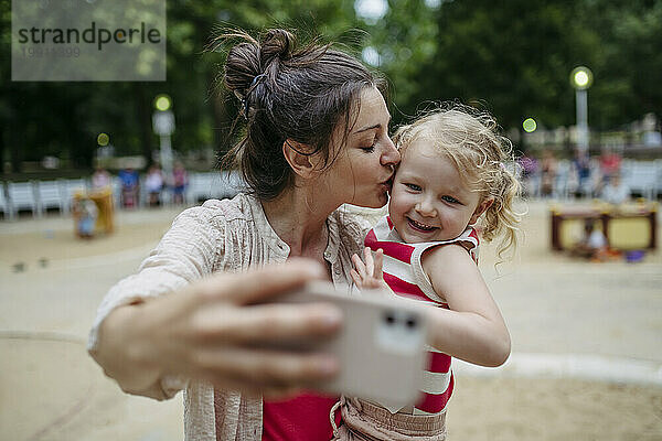 Kleines Kleinkindmädchen und Mutter haben Spaß auf dem Spielplatz und machen Selfies mit dem Smartphone