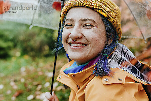 Lächelnde Frau mit Strickmütze und Regenschirm im Herbstpark