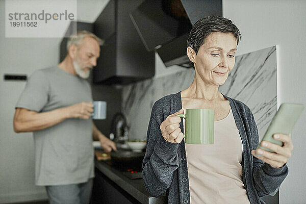 Frau benutzt Smartphone  während ihr Mann im Hintergrund das Mittagessen kocht