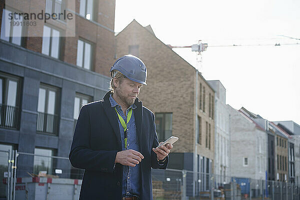 Architekt trägt Schutzhelm und benutzt Mobiltelefon in der Nähe von Gebäuden
