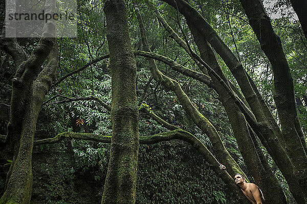 Shirtless man leaning on tree trunk in forest