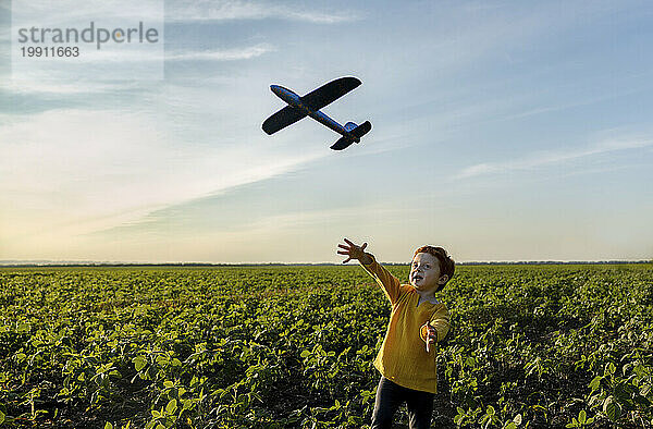 Sorgloser Junge spielt mit Spielzeugflugzeug auf landwirtschaftlichem Feld unter Himmel
