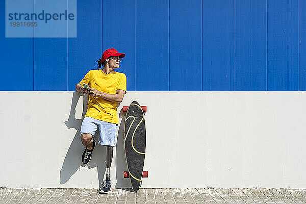 Junger Mann mit Behinderung hält Smartphone in der Hand und steht an sonnigem Tag neben Skateboard