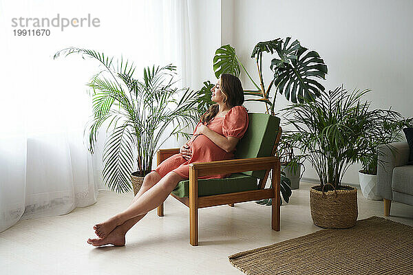 Schwangere Frau entspannt sich zu Hause auf einem Sessel neben Pflanzen