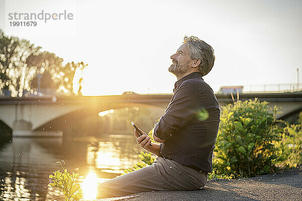 Lächelnder älterer Mann sitzt bei Sonnenuntergang mit Mobiltelefon am See