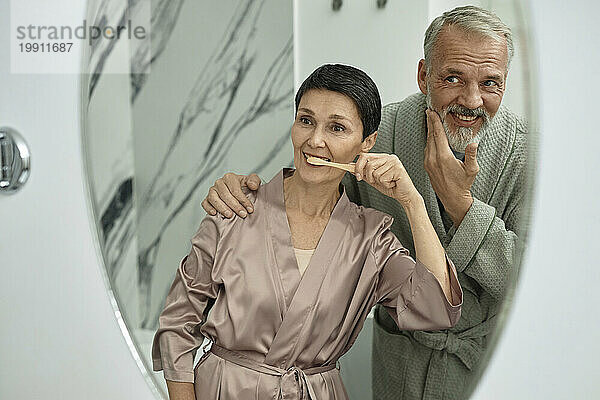Mann kuschelt seine Frau und putzt vor dem Spiegel die Zähne