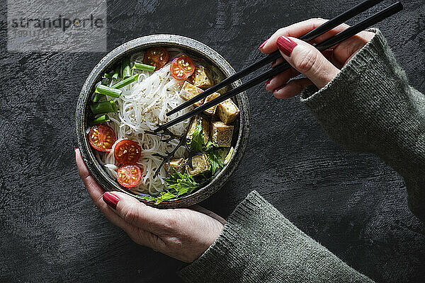 Hands of woman eating bowl of vegan Tom kha kai soup with tofu  tomatoes  salad  rice noodles  sesame seeds and scallion