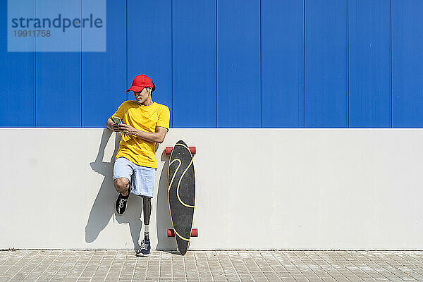 Mann mit Behinderung benutzt Smartphone an sonnigem Tag in der Nähe von Skateboard