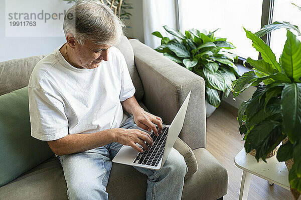 Älterer Mann sitzt zu Hause auf dem Sofa und benutzt Laptop