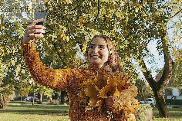 Glückliche Frau hält einen Haufen Ahornblätter in der Hand und macht ein Selfie im Herbstpark