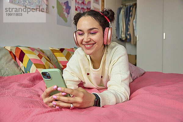 Lächelnde junge Frau  die im Schlafzimmer Musik über Kopfhörer hört und telefoniert