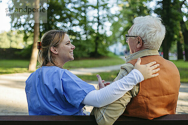 Fröhlicher Mitarbeiter im Gesundheitswesen unterstützt und tröstet den älteren Mann auf der Bank im Park