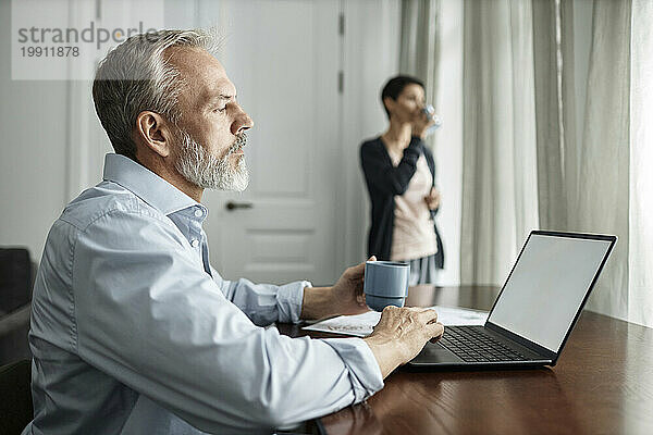Mann benutzt Laptop und trinkt Tee  während er Zeit zu Hause mit seiner Frau verbringt