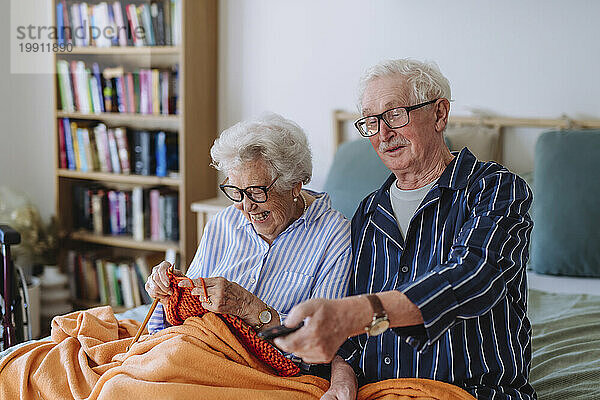 Glückliche Seniorin strickt mit Mann  der die Fernbedienung in der Hand hält und zu Hause fernsieht