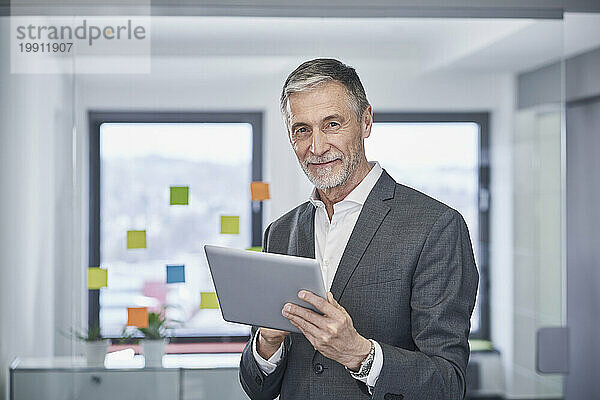 Lächelnder älterer Geschäftsmann steht mit Tablet-PC im Büro