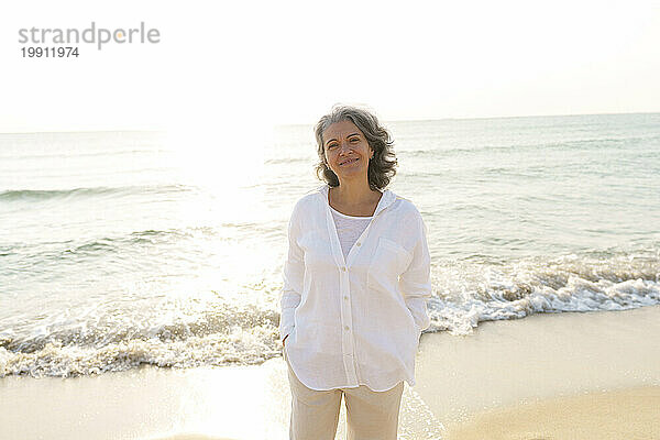 Lächelnde reife Frau mit den Händen in den Taschen  die am Strand steht