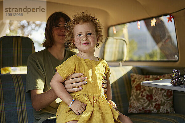 Lächelndes unschuldiges Mädchen  das im Wohnwagen auf dem Schoß seines Bruders sitzt