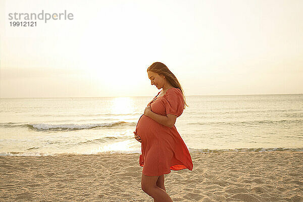 Glückliche schwangere Frau mit den Händen auf dem Bauch  die am Strand vor dem Meer steht