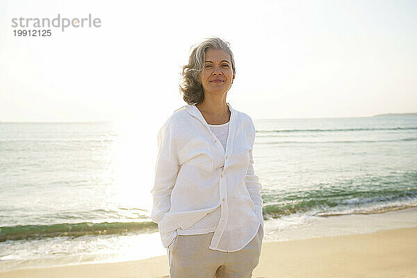 Lächelnde reife Frau  die an einem sonnigen Tag vor dem Meer am Strand steht
