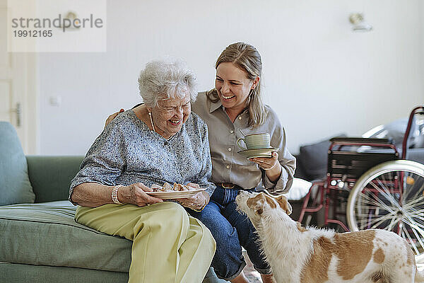 Glückliche ältere Frau hält einen Teller mit einem Gesundheitshelfer  der zu Hause auf dem Sofa neben dem Hund sitzt