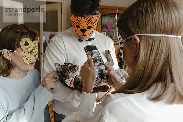 Familie trägt Tiermasken und fotografiert Katze per Smartphone