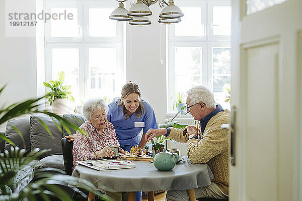 Lächelnder Mitarbeiter im Gesundheitswesen mit älterem Paar  das am Tisch Schach spielt