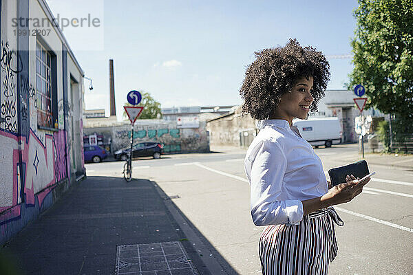 Smiling young businesswoman holding smart phone standing at roadside in city