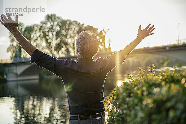 Senior man standing with arms raised near lake at sunset