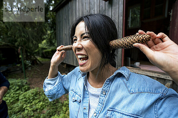 Fröhliche Frau hält Tannenzapfen über die Ohren vor einer Holzhütte