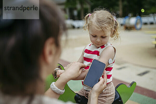 Mutter mit Smartphone schaut auf kleines Mädchen mit einem kontinuierlichen Glukosemonitor-Sensor auf ihrer Schulter
