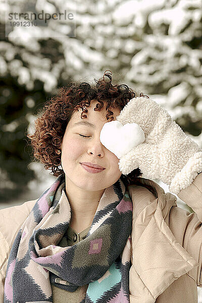 Lächelnde Frau  die im Winter ihr Auge mit herzförmigem Schnee bedeckt