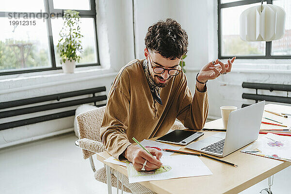 Junger Illustrator macht Design auf Papier mit Laptop am Schreibtisch im Büro