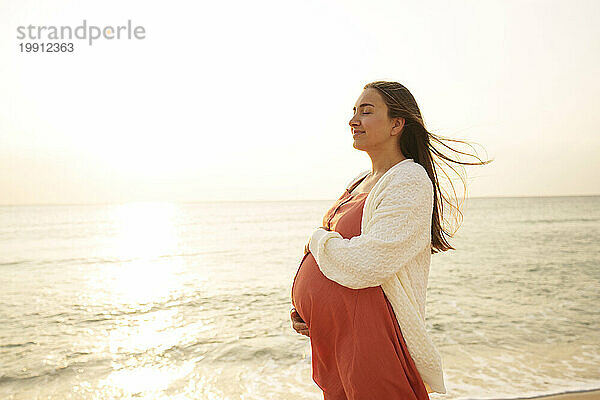 Lächelnde schwangere Frau steht mit geschlossenen Augen am Strand