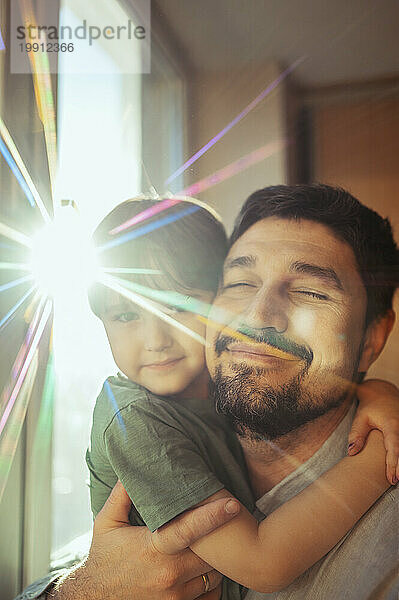 Smiling father with eyes closed embracing son near window