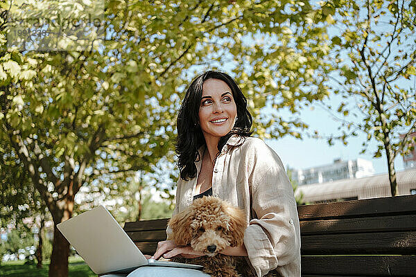 Smiling freelancer sitting with laptop and poodle dog on park bench