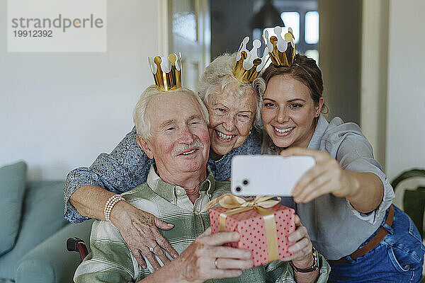 Fröhlicher Mitarbeiter im Gesundheitswesen  der ein Selfie mit einem älteren Paar macht und zu Hause Geburtstag feiert