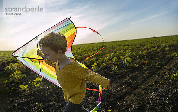 Happy boy holding kite and running in agricultural field under sky
