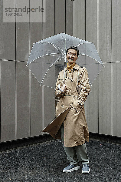Glückliche Frau in beigem Regenmantel hält Regenschirm in der Hand  während sie draußen geht und in die Kamera blickt