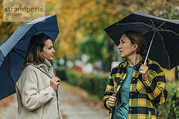 Mother with daughter holding umbrellas and talking in autumn park