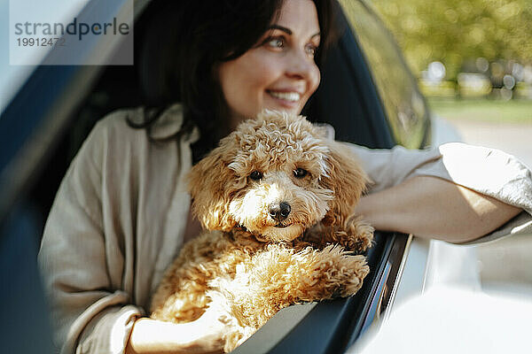 Smiling woman holding poodle dog and leaning out of window in car