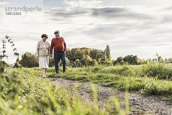 Älteres Paar hält Händchen und geht unter bewölktem Himmel auf Fußweg